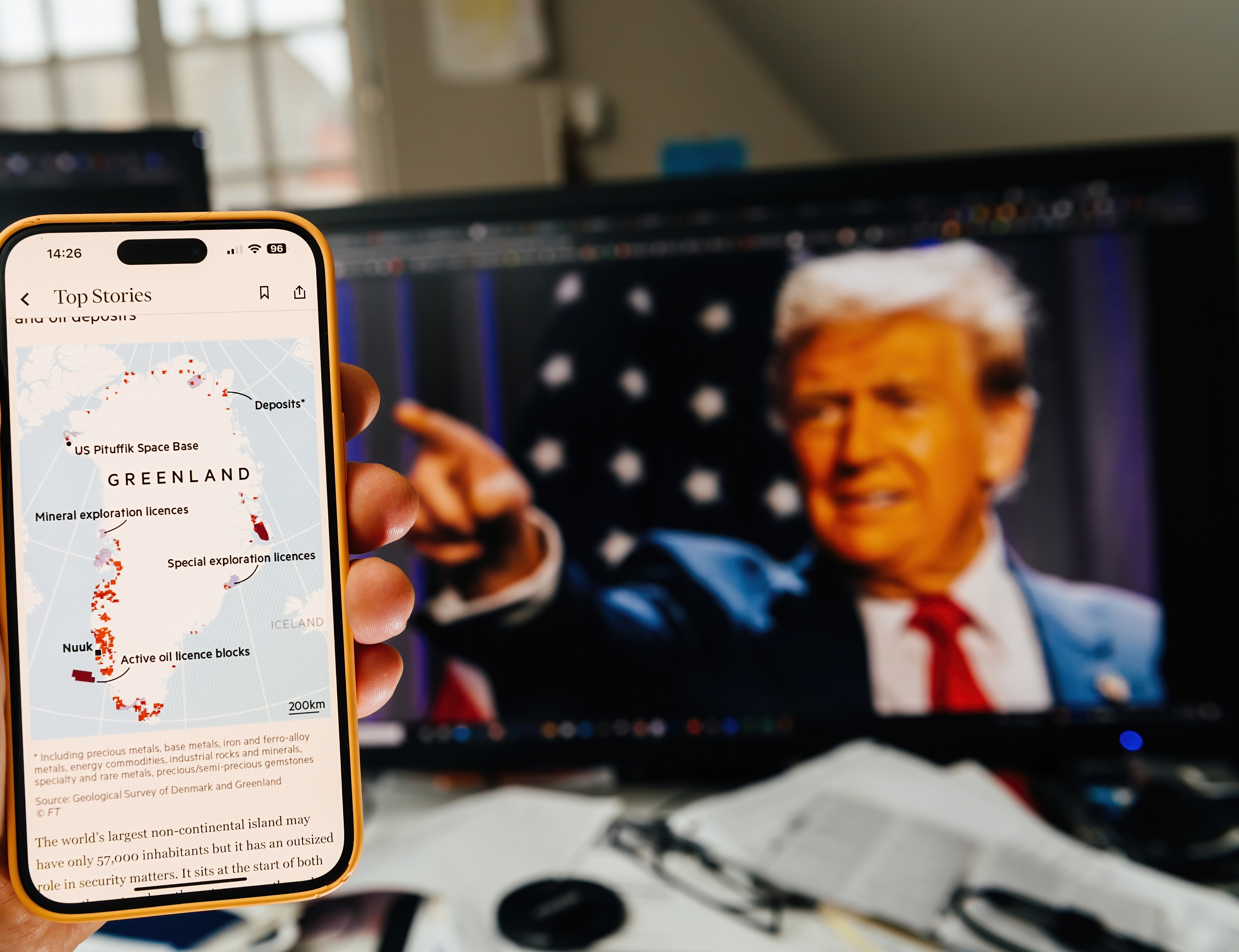 Une main tenant un smartphone affichant une carte des ressources du Groenland avec des détails d'exploration, tandis que deux moniteurs montrent Donald Trump pointant du doigt et une carte du Groenland.